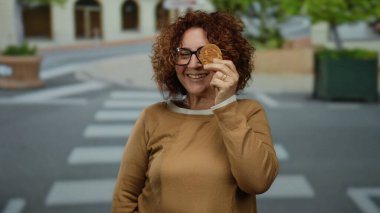 Şehir caddesinde kurabiyelerden hoşlanan bir kadın neşeli bir ifade sergiliyor, kıvırcık esmer saçlar takıyor, gözlük takıyor, şehir arkaplanını rahat ve neşeli bir tavırla harmanlıyor..