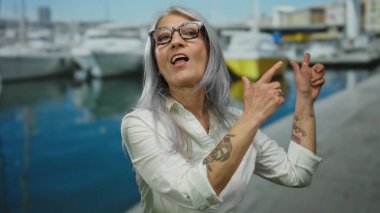 Limanda teknelerin yanında gri saçlı bir kadın, gözlüklü ve beyaz gömlekli, canlı deniz manzaralı bir dövme sergiliyor..