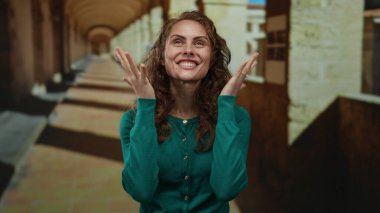 Üniversite koridorunda gülümseyen, neşeli duygular ve etkileyici jestler sergileyen bir kadın eski akademik ortamda yeşil bir kazak giyerken tuğla detaylarıyla.