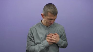 Gri gömlekli beyaz adam, mor izole edilmiş bir arka plana karşı duygusal bir ifadeyle göğsünü tutuyor..
