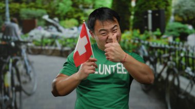 Şehir sokağında elinde İsviçre bayrağı tutan genç bir Asyalı yeşil gönüllü gömleğiyle pozitif bir ifade sergiliyor.