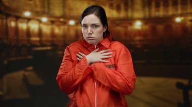 Turuncu bir hapishane tulumu giyen kadın, loş bir Katolik kilisesinin içinde göğsüne dayanmış kasvetli bir ifadeyle titriyor..