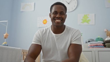 Sıcak bir yatak odasında bebek beşiği ve yumuşak dekoruyla otururken içten içe gülümseyen genç adam mutluluk ve rahatlık saçıyor..