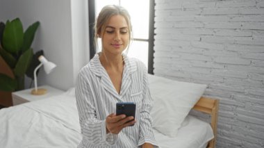Sıcak yatak odasında gülümseyen, elinde akıllı telefon, pijamaları olan, parlak ev iç kısımlarında yatağında oturan, arka planında tuğla duvar ve bitki olan bir kadın.