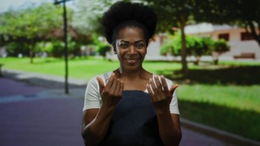 Önlüklü kadın parkta jest yapıyor ağaçlar, çimenler ve patika ile, davetkar, sıcak ifade sergiliyor, açık havayı aydınlatıyor, doğal, huzurlu çevreyi..