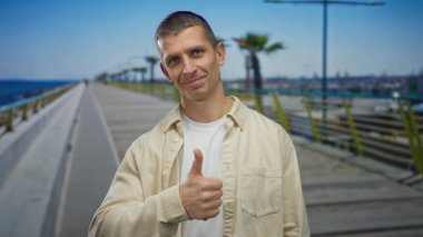 Gülümseyen adam, arka planda okyanus ve palmiye ağaçları olan deniz kenarındaki gezinti güvertesinde rahat bir açık hava yaşam tarzı ve pozitif bir ifade sergiliyor..