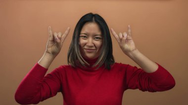 Kırmızı kazaklı kadın kahverengi duvara karşı kaya hareketi yapıyor, neşe ve enerji ifade ediyor, canlı bir ifade ve gençlik coşkusu sergiliyor..