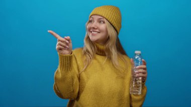 Su şişesiyle gülümseyen, sarı süveter giyen ve bereli bir kadın, izole edilmiş mavi bir arkaplanı işaret ediyor, hidrasyon ve pozitifliği gösteriyor..