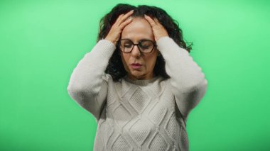 Kıvırcık saçlı kadın yeşil arka planda acı içinde başını tutuyor. Stres ve rahatsızlık anlarını yakalıyor. Sıradan giyinmiş, orta yaş zorluklarını canlı bir şekilde temsil ediyor..