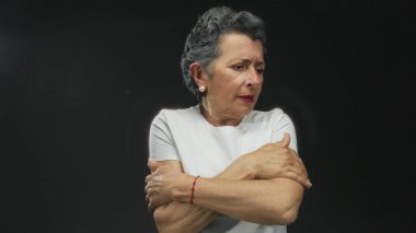 Yaşlı bir kadın, gri saçlı, siyah bir arka planda soğuk görünüyor, kollarını tutuyor ve beyaz bir gömlek giyiyor, soğuk bir rahatsızlık veriyor..