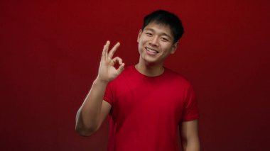 Kırmızı tişörtlü genç Çinli adam, izole edilmiş kırmızı arka planda onay ve güvene karşı tamam işareti gösteriyor.
