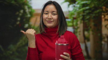 Kırmızı kazaklı kadın şehir caddesinde duruyor, gülümsüyor ve içki şişesi tutuyor, açık havada şehir ortamını işaret ediyor..