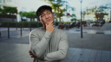 Gri kazaklı genç adam, bulanık şehir arka planı ve yemyeşil ile çevrili hareketli bir şehir caddesinde düşünceli bir şekilde poz veriyor..