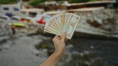 Deniz kenarındaki bulanık plaj geçmişiyle elinde kara para tutan bir adam açık hava ortamında finansal işlem kavramını resmediyor..