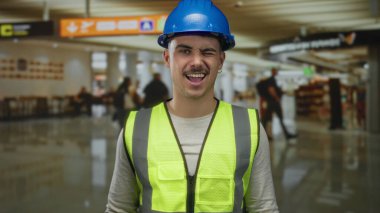 Yansıtıcı yelek giyen, şapkalı, dilini dışarı çıkaran ve arka planı bulanık ve yön işaretleri olan bir havaalanı terminalinde göz kırpan genç bir İspanyol..