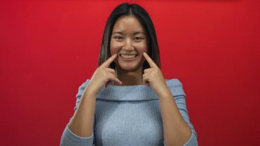 Canlı kırmızı bir duvarın önünde parmakları yanaklarında gülümseyen bir kadın, açık mavi süveter giyen, izole edilmiş arka plan, neşe ve pozitiflik gösteren genç bir kadın..