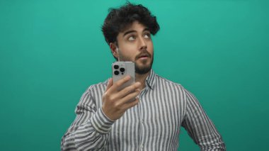 Sakallı ve çizgili gömlekli genç adam canlı yeşil arka planda akıllı telefon tutuyor, meraklı ve nişanlı görünüyor, modern teknoloji ve İspanyol kimliğini öneriyor..