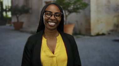 Sarı bir bluz ve gözlük takan bir kadın, bir şehir sokağında dikiliyor, mutluluğu ve kendine güveni iletiyor..