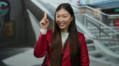Uzun saçlı, kırmızı ceketli, alışveriş merkezinde gülümseyen bir kadın, parmağını kaldırıyor, modern, Asyalı, şehir hayatı tarzını gözler önüne seriyor, parlak iç görünüşlü ve hareketli bir alışveriş ortamı var..