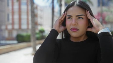 Sokakta siyah bluzlu bir kadın, şehir ortamında parlak güneş ışığı altında gözleri kapalı tapınaklara baskı yaparken stresli görünüyor..