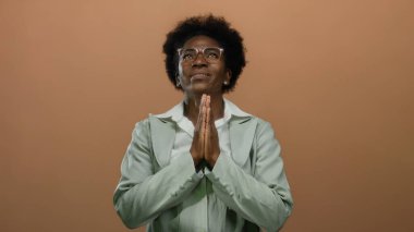 Elleri sımsıkı bağlı, gözlüklü bir kadın, tenha kahverengi bir arka plana karşı duruyor, huzurlu bir duygu ve inanç iletiyor, huzurlu, yansıtıcı bir ortama.