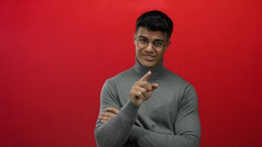 Genç İspanyol bir adam, parmaklarıyla kırmızı bir arkaplan karşısında hayır işareti yapıyor. Gri balıkçı yaka bir kazak içinde cazibe ve kendine güven yayıyor. Çekici bir gülümseme sergiliyor..