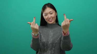 Gri süveter giymiş, orta parmaklı, canlı yeşil bir arkaplan karşısında gülümseyen bir kadın cesur bir ifade ve kendine güvenen bir tavır takınıyor, günlük giysiler sergiliyor..