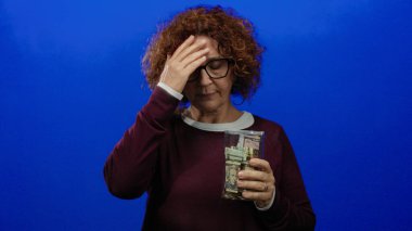 Dolarla dolu cam bir kavanozu tutan kadın, soyutlanmış bir mavi arka planda endişeli görünüyor..