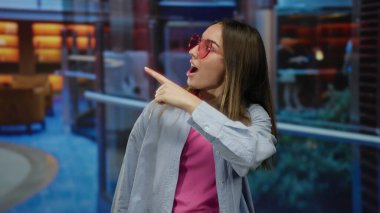 Sarı saçlı, pembe güneş gözlüğü takan ve şık dekorlu, mavi halılı modern bir otel ortamını gösteren genç bir kadın..