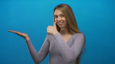 İzole edilmiş mavi bir arkaplan karşısında el kol hareketi yapan genç bir kadın, gelişigüzel bir ortamda pozitiflik ve güven gösteriyor, ifadesini ve vücut dilini vurguluyor..