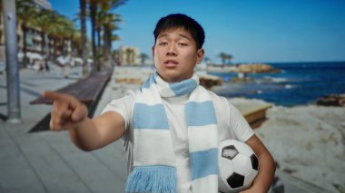 Futbol toplu genç adam deniz kenarındaki bir güvertede duruyor, çizgili bir eşarp takıyor, güneşli plaj gününün tadını çıkarıyor ve etrafında palmiye ağaçları var..