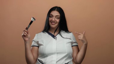 Genç, kıvrımlı bir kadın yüzünde bir gülümsemeyle izole edilmiş bir kahverengi duvar arkaplanına karşı makyaj fırçası tutuyor, özgüven ve güzellik sergiliyor..