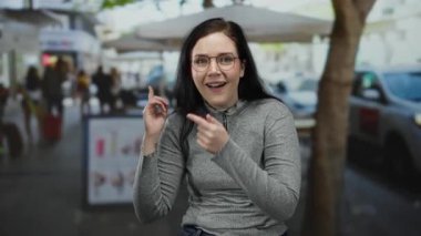 Arka planda bulanık insanlar ve arabalarla dolu canlı bir sokakta heyecanlı bir şekilde el kol hareketi yapan bir kadın, dışarıda neşe ve sürpriz taşıyor..