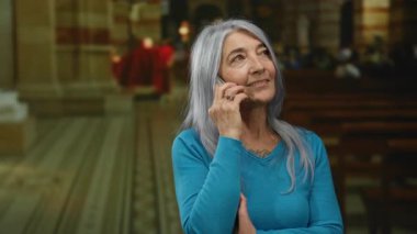 Kilisenin içinde telefonla konuşan gri saçlı yaşlı bir kadın, mavi kazak giyiyor, sıcak ışıklandırmayla ruhsal ortamda derin düşüncelere dalmayı öneriyor..