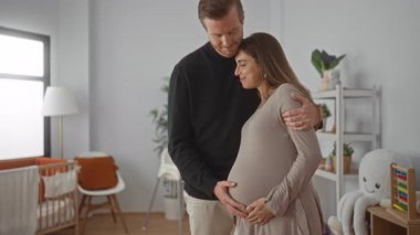 Hamile kadın ve erkek bebek odasında neşeyle kucaklaşıyorlar. Beşik, kitap ve oyuncaklarla. Sevgi ve beklentiyle dolu huzurlu bir ev ortamı yaratıyorlar..