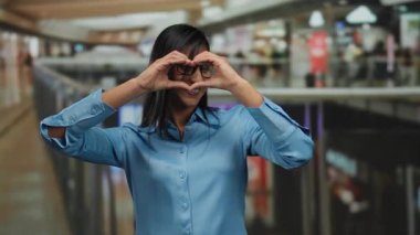 Kapalı bir alışveriş merkezinde, elleri mavi gömlek ve gözlük takarak kalp şeklinde bir kadın, pozitif ve dostane bir atmosfer yaratıyor..
