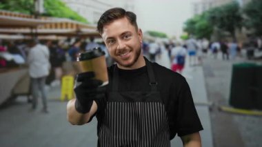 Önlüklü genç adam açık hava pazarında elinde hazır kahveyle şehir arka planında hareketli gülümsüyor ve sokak ortamında içki servisi yapan bir garson gibi gülümsüyor..