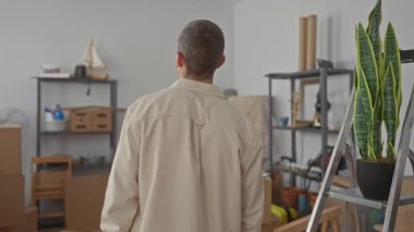 Yeni dairenin oturma odasında duran adam gülümseyen ve rahatlayan kutularla ve raflarla çevrili.