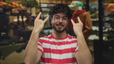 Sakallı, çizgili tişörtlü genç adam, parlak bir geçmişi olan bir giyim mağazasında taş gibi jest yapıyor.
