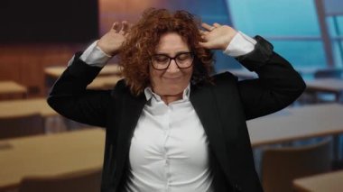 Ahşap masalı ve modern tasarımlı, gözlüklü ve takım elbiseli orta yaşlı esmer bir kadın, bir sınıfta uyukluyor ve el kol hareketi yapıyor..