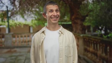 Ağaçlarla çevrili arka planda gülümseyen bir adam, tasasız, rahat, samimi bir an yakalıyor. Parlak ve renkli bir ortamda pozitif, rahat bir atmosfer tasvir ediyor..