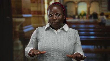 Kilise ortamında gözlüklü ve gri kazaklı Afro-Amerikalı bir kadın, sakin görünüyor, elleri açık, geleneksel kilise sıraları ve hafif aydınlatma ile çevrili..
