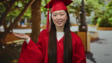 Kırmızı mezuniyet elbisesi ve şapkalı kadın yemyeşil bir parkta duruyor. Elleriyle, başarıyı ve kutlamaları sembolize ederken neşeli bir ifadeyle ileriye bakıyor..