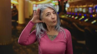 Kumarhanede gri saçlı bir kadın kumar makinelerinin önünde el kol hareketi yapıyor. Canlı bir kumar ortamında merak ve tefekkür taşıyor..