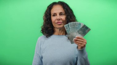 Orta yaşlı bir kadın, izole edilmiş yeşil bir duvara karşı Brezilya banknotları tutuyor. Finansal başarı ve pozitiflik sergiliyor..