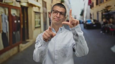 Gözlüklü ve çizgili tişörtlü bir adam Avrupa caddesinde ayakta duruyor. Kaybedenin işareti ile kameraya neşeli bir ifadeyle bakıyor..