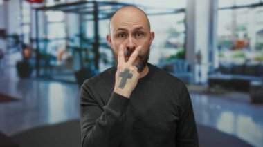 Oteldeki kel adam, parmakları gözlerine yakın, çapraz dövmeli, koyu renk süveterli, kapalı alanda bulanık duran, dikkatli bir jest yapıyor..