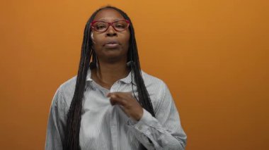 Gözlüklü ve uzun saçlı bir kadın turuncu bir duvara dayanıyor. Çizgili bir gömlek giyiyor. Rahat ve huzurlu görünüyor..