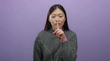 Sessizliği işaret eden bir kadın, parmak dudaklarında, soyutlanmış mor bir arka plan karşısında duruyor, sakin bir tavırla güzelliğini ve Asyalı etnik kökenini vurguluyor..