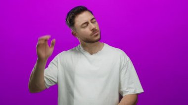 Sakallı genç adam, boynu rahatsız edici bir şekilde arka planda, izole edilmiş pembe duvara yaslanmış, rahatsız edici bir anı yakalıyor ve günlük beyaz giysiler içinde iç gözlem yapıyor..